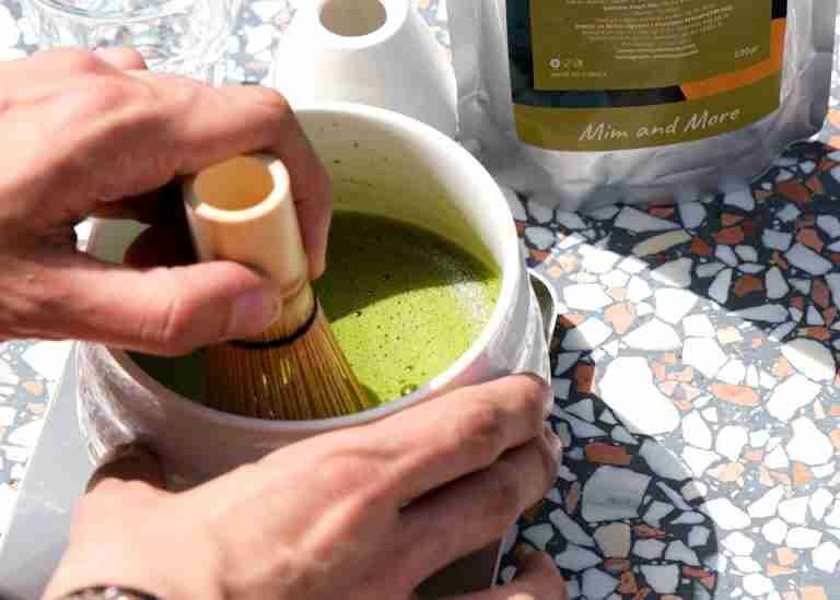 A person whisking vibrant green matcha tea in white bowl using a bamboo whisk on a sunny terrazzo table.