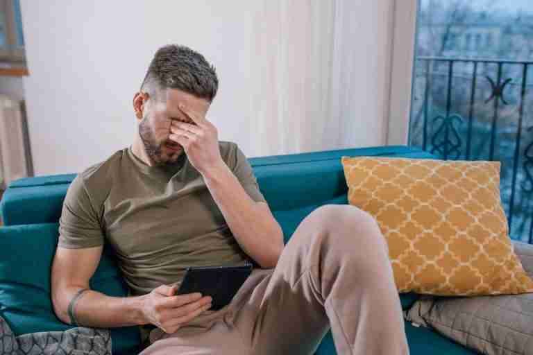 A man sits on a sofa holding a tablet, visibly stressed and covering his face with one hand.