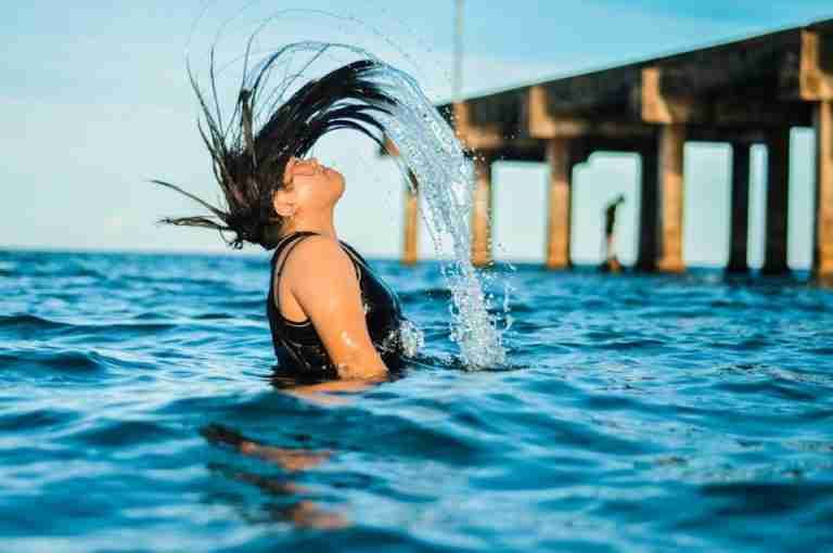 A woman flips her hair in the ocean near a pier on a sunny day, capturing a fun summer moment.