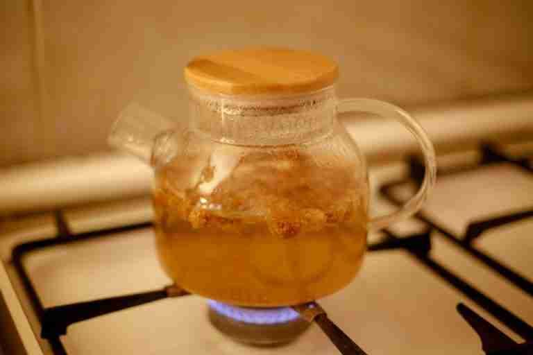 A glass teapot with bamboo lid brewing herbal tea on a gas stove indoors.