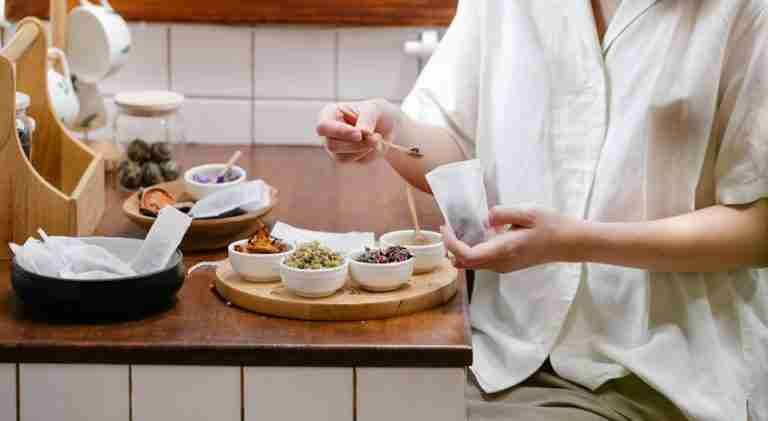 A person in a white shirt preparing herbal tea with assorted dried flowers and herbs on a wooden tray.