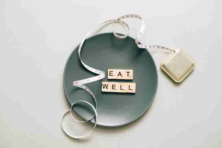 Flat lay of scrabble tiles spelling 'eat well' on a plate with a tape measure, promoting healthy eating.