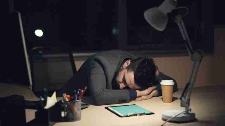 Man in office suit sleeping on desk with coffee and tablet nearby, symbolizing work fatigue.