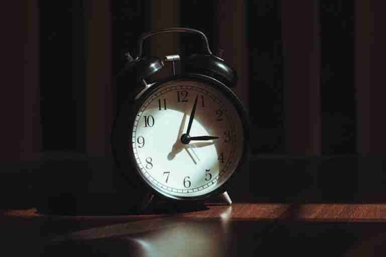 A classic black analog alarm clock illuminated by warm light and artistic shadows.