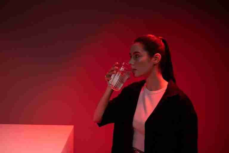 A fashionable woman drinks water in a red-lit room, creating a dramatic ambiance.