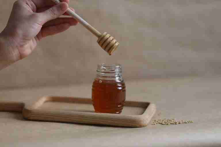 A hand holding a honey dipper over a jar on a wooden tray, showcasing the golden liquid.