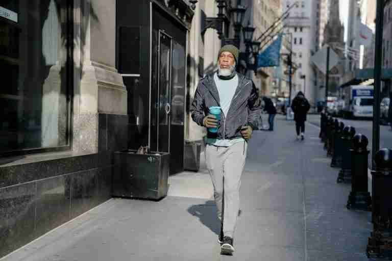 Full body of confident bearded African American male in activewear and warm gloves and hat walking along sidewalk with fitness bottle for water in sunny morning