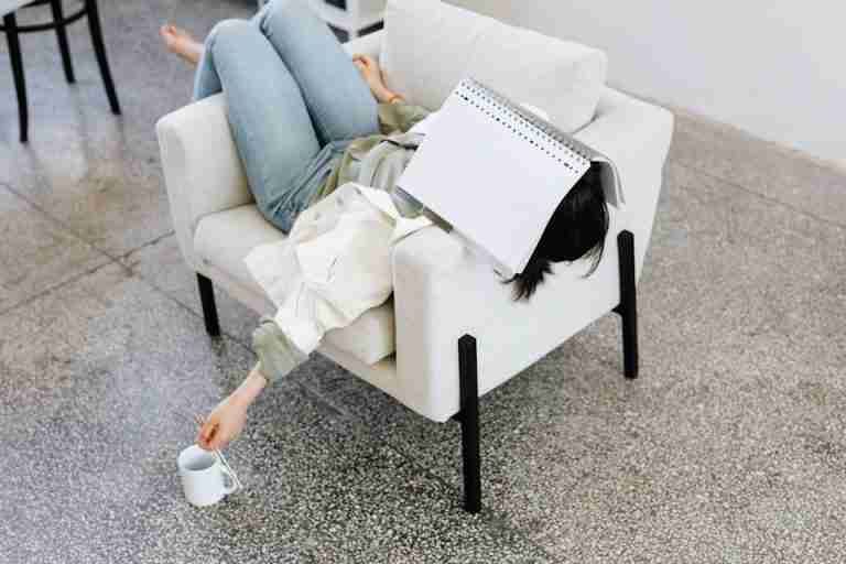 Woman lying on an armchair with a book on face, holding a coffee. Casual and relaxing atmosphere.