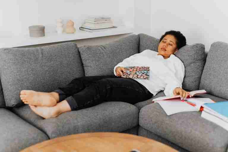 A young woman resting on a cozy grey sofa with open books around her, appearing tired after studying.