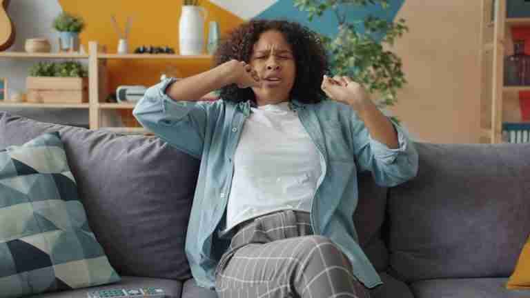 Woman yawning on a living room sofa, wearing comfortable casual clothes.