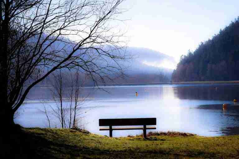 Serene landscape of a lake with an empty bench and misty hills in the background.