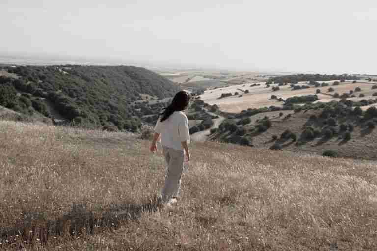 A woman walking peacefully on a hillside overlooking a serene valley and vast fields.