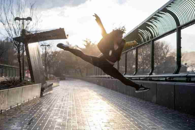 A dancer leaps gracefully on a rainy day, backlit by the setting sun in an urban environment.