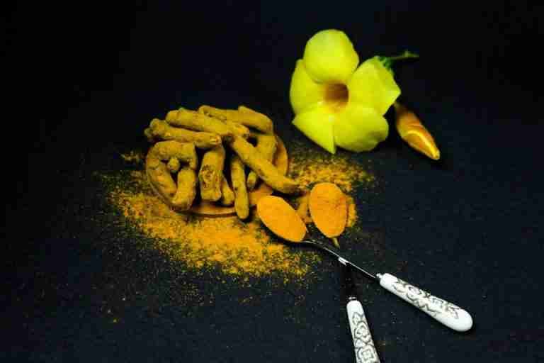 Still life of turmeric powder and root with a yellow flower on a dark surface.