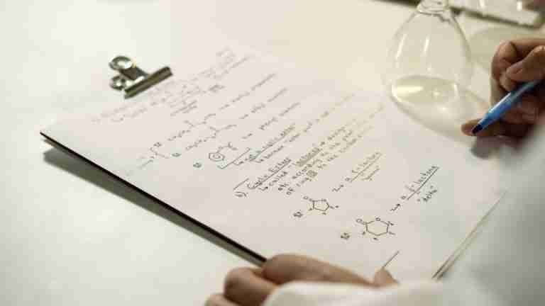 A close-up view of handwritten chemical formulas on a clipboard in a lab setting.