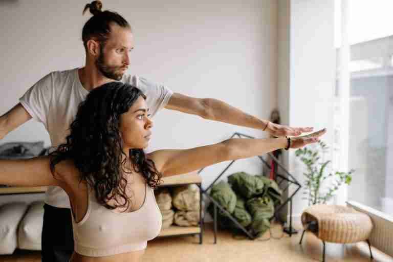 A yoga instructor helps a woman with her Warrior Pose in a serene indoor studio.