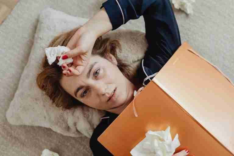 A woman lies on a pillow, holding crumpled tissues, conveying a sense of sadness or exhaustion.