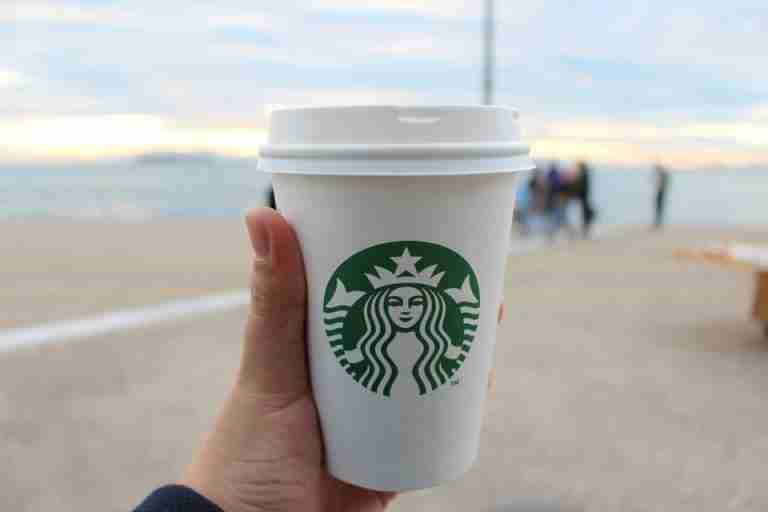 A hand holds a Starbucks coffee cup by the seaside, capturing a serene outdoor moment.