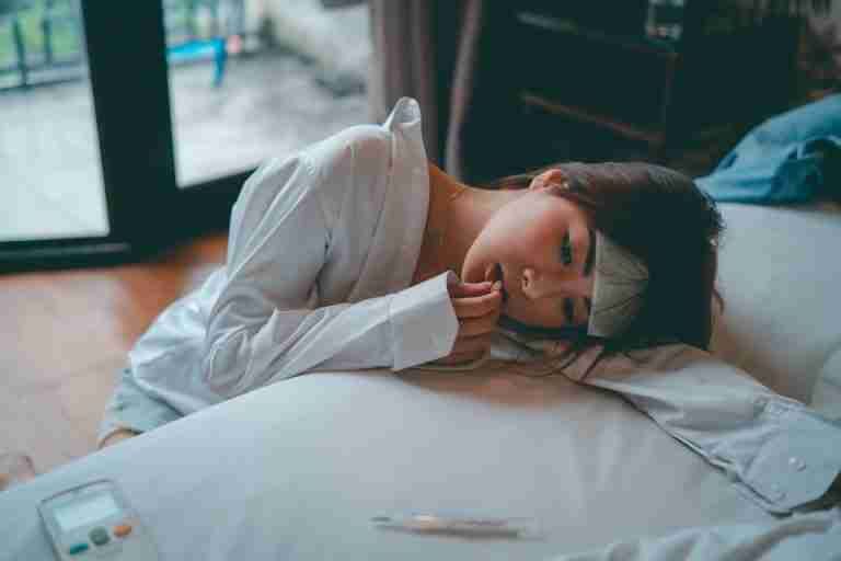 A young woman lies in bed, appearing unwell, taking medicine to alleviate symptoms.