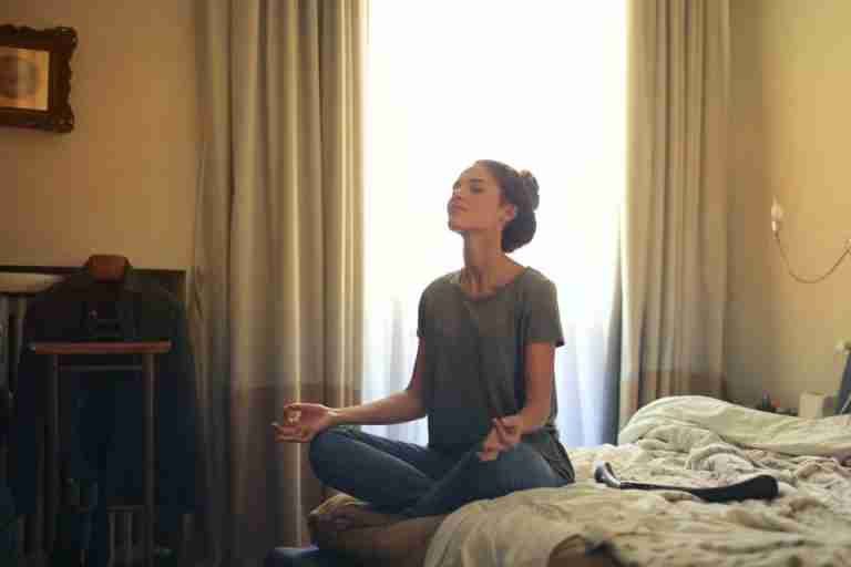 Adult woman practicing meditation on her bed surrounded by a calm bedroom atmosphere.