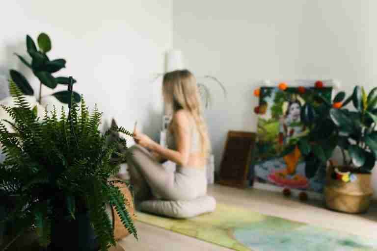 A woman practices meditation indoors surrounded by plants, promoting wellness and relaxation.