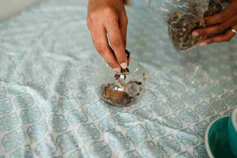Close-up of hands filling a teapot with dried herbal leaves on a patterned cloth surface.