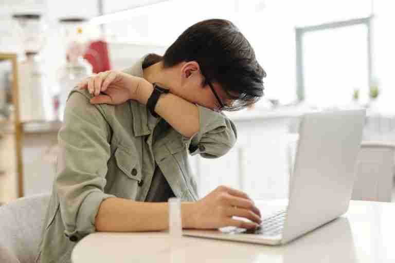 A man sneezes into his elbow while using a laptop, depicting common cold symptoms in a modern workspace.